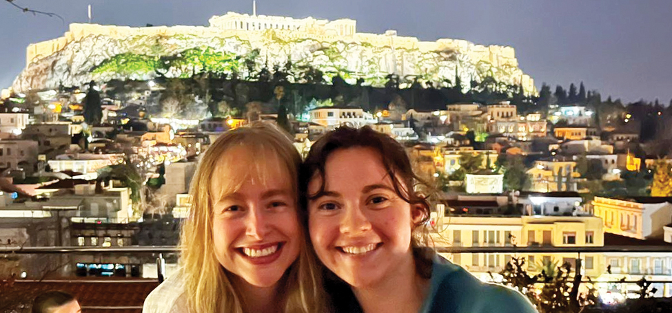 Two women pose, heads together, with Athens in the background