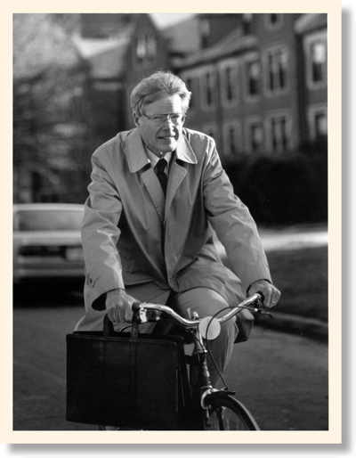 George Drake in a light dress coat riding a bike while holding his briefcase in his right hand 