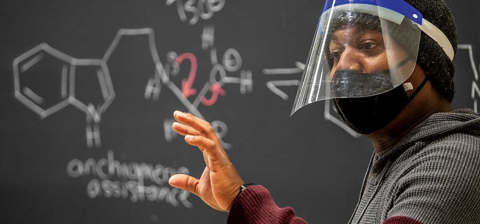 Erick Leggans in a mask and face shield during the pandemic speaking from in front of a blackboard with molecular diagrams on the board behinid