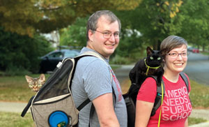 Two people with leashed cats, one of which is in a backpack and the other on a shoulder