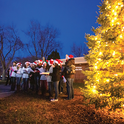 group of people in santa hats sing together a tree with lights to the right