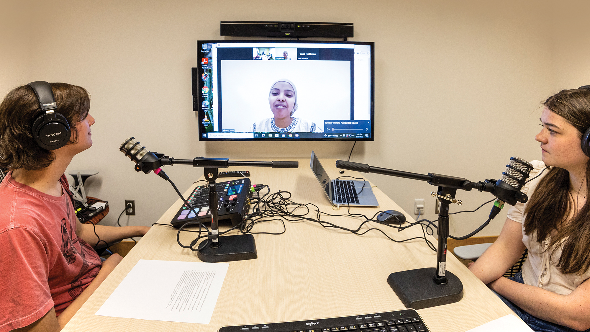 2 people with head- and microphones are facing a monitor that displays an online meeting