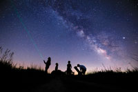 Grass and students silhouetted against a sky bright wth stars with a green beam from a flashlight shiing towards the sky