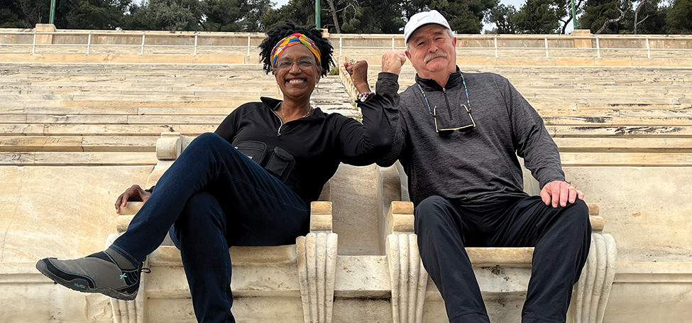 Kesho Scott and Will Freeman sit in side-by-side stone sculpted throne-like chairs in a stone stadium, lifted arms touching