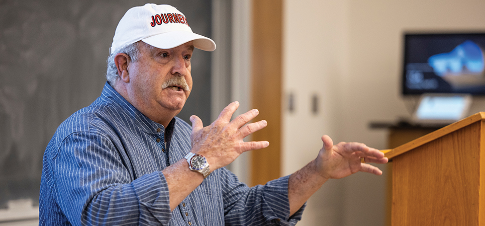 Will, wearing a white ball cap with Journeys stitched in red, gestures while speaking to the class