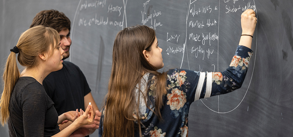 A student writes on a chalkboard while another stands ready, chalk in hand, and a third watches