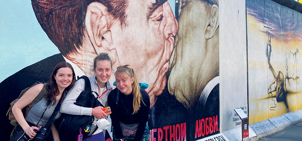 Three students pose in front of a mural on the Berlin Wall