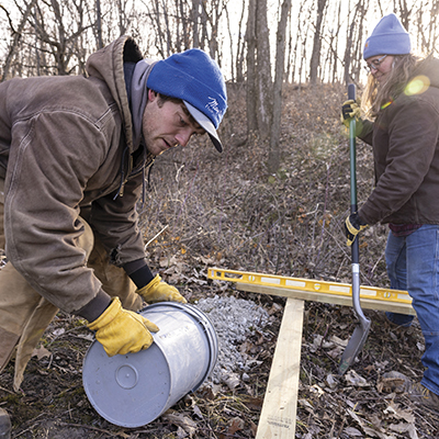 Jake Hill in a brown coat and blue cap upends a 5-gallon bucket of gravel while Emily Klein in a blue hat stands ready with as shovel