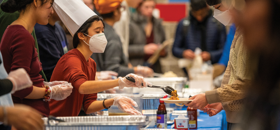A student in chef's hats and gloves serve food to a guest holding out their plate across a table