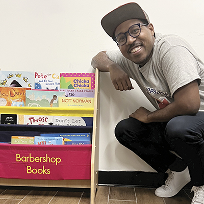 Alvin Irby crouches next to a colorful book holder with Barbarshop Books in bright yellow on the front