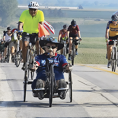 Scott Porter rides his handcycle with bicyclists. He wears a large hat and dark glasses with a nose guard
