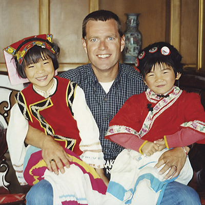 Scott Raecker holds two young Chinese girls in traditional dress on his lap