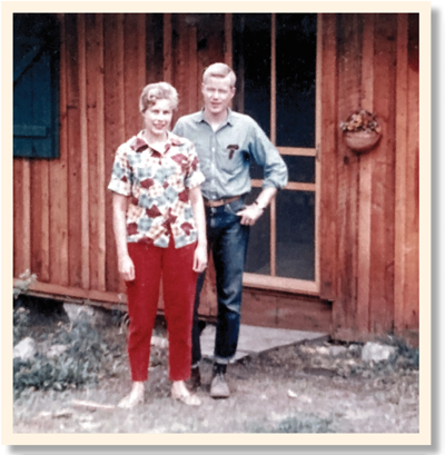 Man and woman stand in front of the screen door of a wooden building