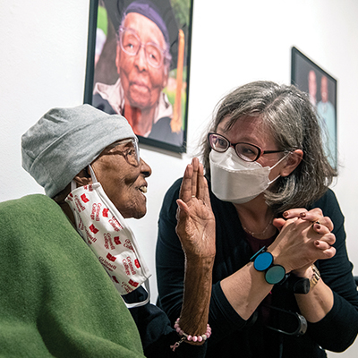 Older Black woman holds hand up while talking to a white woman in a white mask a painting of the Black woman in academic garb hangs behind them
