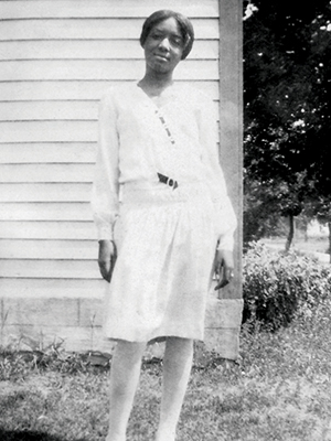 A Black girl in a white dress and tights stands outside of a house