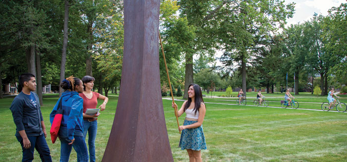 Students at the base of a metal sculpture on campus, one holding a pole