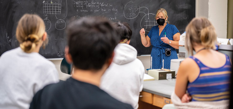 professor in face mask addresses student in lecture room in front of black board