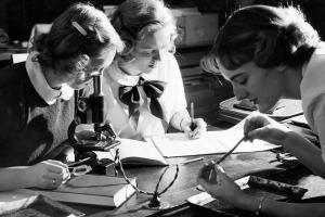 Three women work in a lab, one peering into a manual microscope while another writes i a notebook