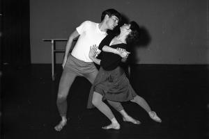 Black and white photo of two dancers, the woman in front, facing the man, with one leg stretched behind her. They are clasping hands
