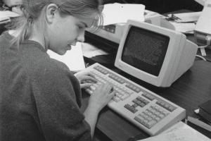Student types on a bulky keyboard in front of a mainframe terminal; a dot-matrix printer in the background.