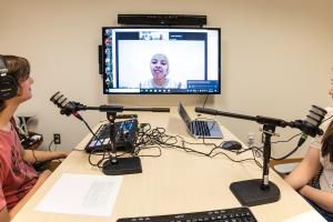 2 people with head- and microphones are facing a monitor that displays an online meeting