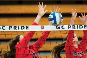 Two players reach for the volleyball behind net printed with GC PRIDE and Honor G